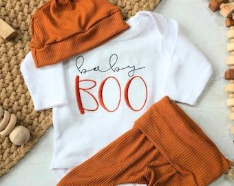 May include: A baby outfit set in orange and white. The set includes a long-sleeved bodysuit with the text "baby BOO" embroidered on the front, matching trousers, and a knotted beanie.