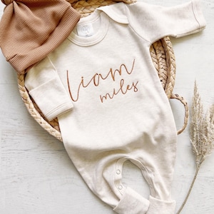 May include: A cream colored long-sleeved baby onesie with the name "Liam Miles" embroidered in brown on the front. The onesie is lying on a woven basket with a brown knitted hat next to it.