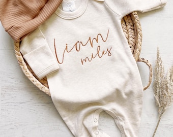 May include: A cream coloured long-sleeved baby onesie with the name "Liam Miles" embroidered in brown on the front. The onesie is lying on a woven basket with a brown knitted hat next to it.