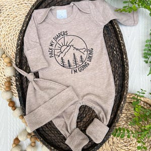 K&ouml;nnte beinhalten: Beige Baby-Set, bestehend aus einem Strampler und einer passenden M&uuml;tze. Der Strampler hat eine schwarze Grafik einer Berglandschaft mit dem Text "PACK MY DIAPERS I'M GOING HIKING". Das Set ist aus weichem Material f&uuml;r Neugeborene.