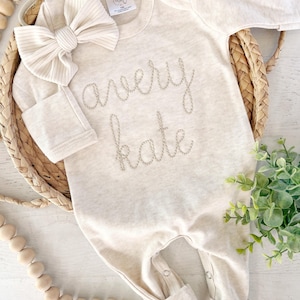 May include: A long-sleeve beige baby bodysuit featuring the embroidered name avery kate in a cursive font on the chest, accompanied by a matching ribbed fabric bow headband. The garment includes a neck label and snap closures at the leg openings.