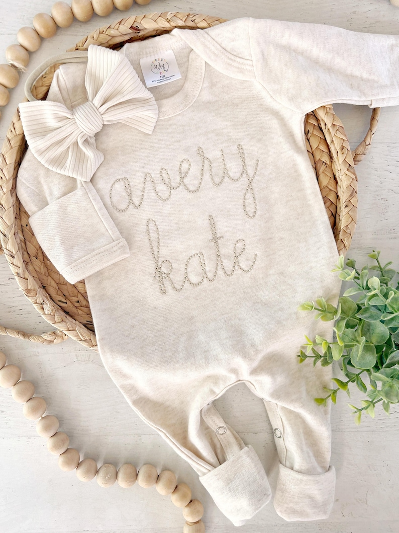 May include: A long-sleeve beige baby bodysuit featuring the embroidered name avery kate in a cursive font on the chest, accompanied by a matching ribbed fabric bow headband. The garment includes a neck label and snap closures at the leg openings.