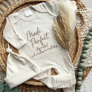 Könnte beinhalten: Beige Baby-Strampler und passendes Mützen-Set mit Knoten. Der Strampler hat den gestickten Text "Made Perfect by a God who makes no mistakes". Das Set wird auf einem geflochtenen Tablett präsentiert.