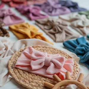 Könnte beinhalten: Eine Kollektion von gerippten Stoffstirnbändern mit einer zentralen Schleife, erhältlich in verschiedenen Unifarben wie Rosa, Senfgelb, Blau, Beige und Grün, ergänzt durch ein Leopardenmuster und einen strukturierten Waffelstrickstoff.