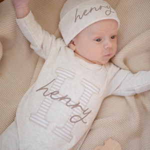 May include: A newborn baby wearing a beige onesie and matching knotted hat, both embroidered with the name "Henry". The onesie features a large letter "H" with striped detailing. A wooden teether is also present.