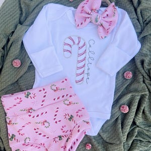 K&ouml;nnte beinhalten: Ein Baby-Outfit mit einem pink-wei&szlig;en Zuckerstangen-Muster, bestehend aus einem wei&szlig;en Langarm-Body, pink-wei&szlig;en Zuckerstangen-Muster-Bl&uuml;mchen und einem rosa Schleifen-Haarband. Der Body ist mit einer gestickten Zuckerstange und dem Namen "Callie" in Kursivschrift verziert.