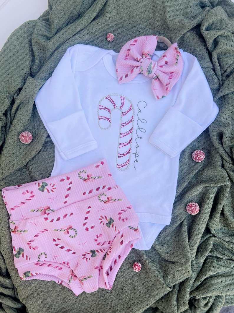 K&ouml;nnte beinhalten: Ein Baby-Outfit mit einem pink-wei&szlig;en Zuckerstangen-Muster, bestehend aus einem wei&szlig;en Langarm-Body, pink-wei&szlig;en Zuckerstangen-Muster-Bl&uuml;mchen und einem rosa Schleifen-Haarband. Der Body ist mit einer gestickten Zuckerstange und dem Namen "Callie" in Kursivschrift verziert.