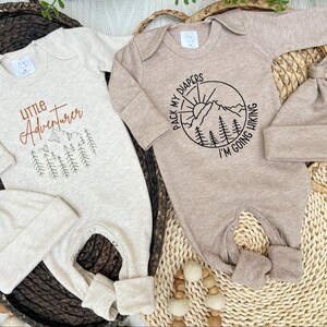 K&ouml;nnte beinhalten: Zwei Baby-Strampler mit passenden M&uuml;tzen. Einer ist beige mit dem Text "Little Adventurer" und einer Berg-Grafik. Der andere ist hellbraun mit einer Berg- und Sonnengrafik und dem Text "Pack my diapers I'm going hiking".
