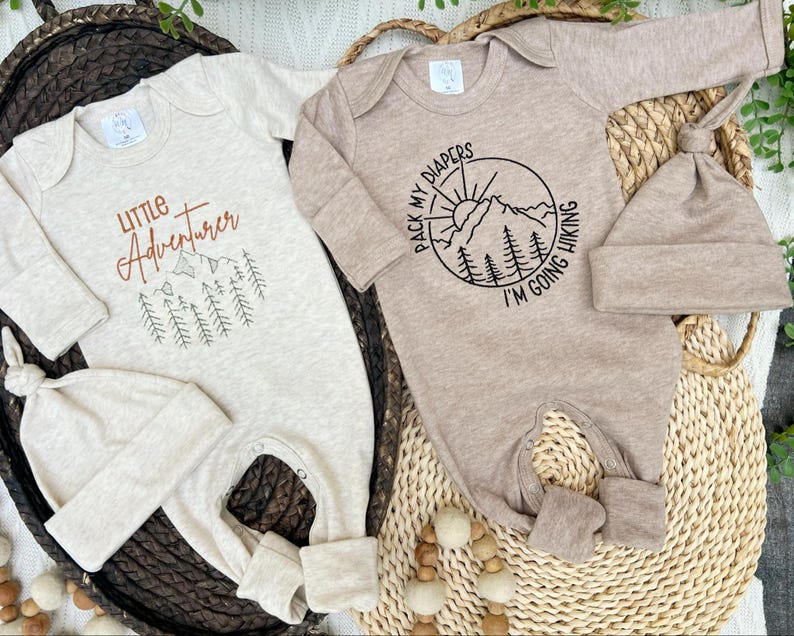 K&ouml;nnte beinhalten: Zwei Baby-Strampler mit passenden M&uuml;tzen. Einer ist beige mit dem Text "Little Adventurer" und einer Berg-Grafik. Der andere ist hellbraun mit einer Berg- und Sonnengrafik und dem Text "Pack my diapers I'm going hiking".