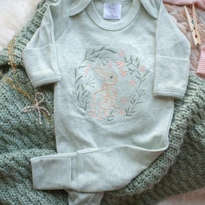 May include: A light green baby onesie with long sleeves and a cute embroidered bunny design. The bunny is surrounded by flowers and leaves. The onesie has a fold-over foot design.