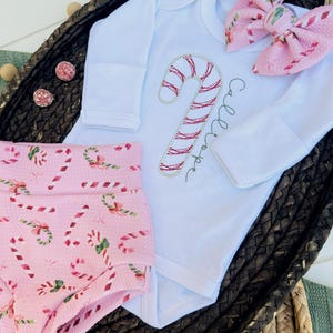 K&ouml;nnte beinhalten: Ein rosa-wei&szlig;es Baby-Outfit mit einem Zuckerstangen-Design. Das Outfit besteht aus einem lang&auml;rmeligen Body mit einer gestickten Zuckerstange und dem Text "Callie Hope" und einer rosa Hose mit einem Zuckerstangen-Druck.