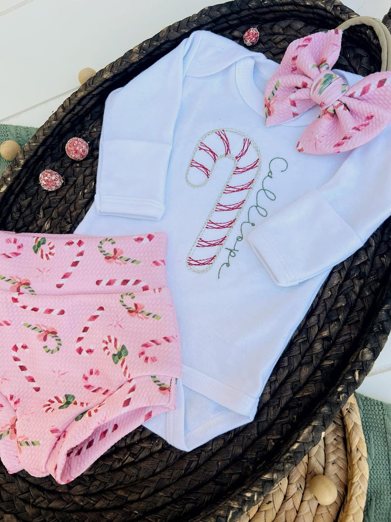 K&ouml;nnte beinhalten: Ein rosa-wei&szlig;es Baby-Outfit mit einem Zuckerstangen-Design. Das Outfit besteht aus einem lang&auml;rmeligen Body mit einer gestickten Zuckerstange und dem Text "Callie Hope" und einer rosa Hose mit einem Zuckerstangen-Druck.