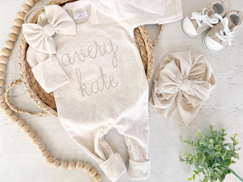May include: A beige long-sleeve baby romper featuring the embroidered name Avery Kate in a cursive font, accompanied by a matching ribbed headband with a large bow, a second ribbed turban-style hat, and a pair of beige canvas sneakers with white laces.