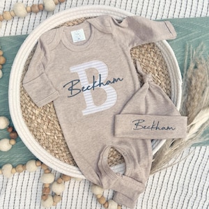 Op de afbeelding: Een beige baby-outfitset, bestaande uit een onesie met lange mouwen en een bijpassende geknoopte muts. De onesie heeft een grote letter "B" met een gestreept ontwerp en de naam "Beckham" in script. De set wordt gepresenteerd op een geweven dienblad.