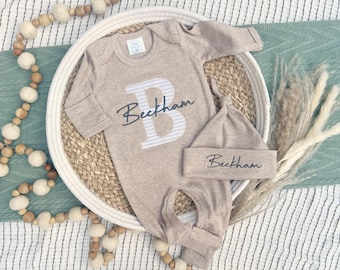 Könnte beinhalten: Ein beiges Baby-Outfit-Set, bestehend aus einem langärmeligen Strampler und einer passenden geknoteten Mütze. Der Strampler hat einen großen Buchstaben "B" mit Streifendesign und den Namen "Beckham" in Schreibschrift. Das Set wird auf einem geflochtenen Tablett präsentiert.