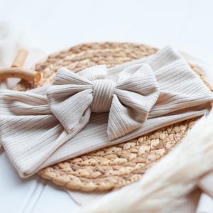 May include: A beige ribbed fabric headband featuring a large, centered bow made from the same material. The headband has a soft, textured appearance with vertical ribbing throughout the fabric, creating a simple and uniform design.