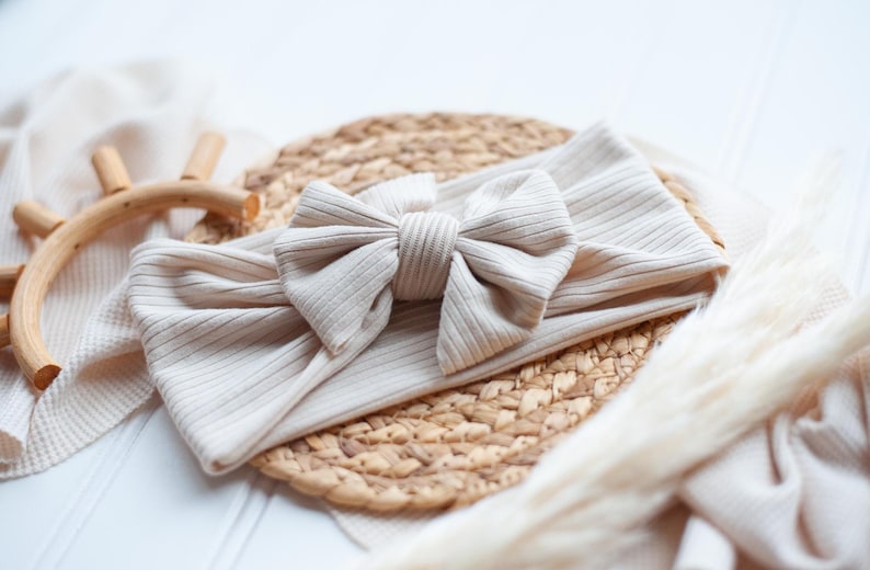 May include: A beige ribbed fabric headband featuring a large, centered bow made from the same material. The headband has a soft, textured appearance with vertical ribbing throughout the fabric, creating a simple and uniform design.