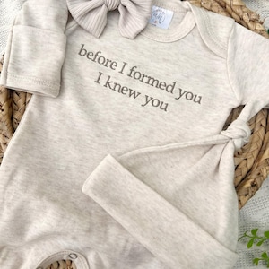 Op de afbeelding: Een beige baby-set met de geborduurde tekst "before I formed you I knew you". Een bijpassende strik-hoofdband en geknoopte muts zijn ook inbegrepen. De outfit is gemaakt van zacht materiaal en is ontworpen voor pasgeborenen.