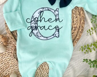 Può includere: Un pagliaccetto per neonati verde menta con maniche lunghe e piedini. Il pagliaccetto presenta il nome "Cohen Grace" in corsivo nero, con la lettera "C" riempita con un disegno floreale. Un fiocco blu navy scuro è posto in alto.