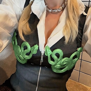 May include: A silver metal necklace and a dark grey vest with a zip. The vest features green snake-shaped decorations. A white collared shirt is worn underneath.