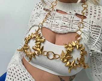 May include: Gold-colored body chain featuring a sunburst design. The chain is composed of circular links and sun-shaped pendants. The model is wearing a white crop top and a white, open-knit sweater. The background is a solid white.