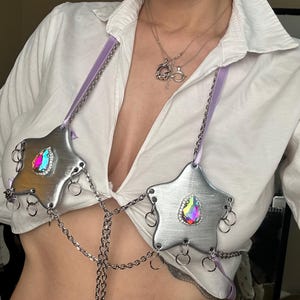 May include: A silver metal body chain with star-shaped breastplates, each with an iridescent gem. The breastplates are linked by a chain and have small rings. Lavender ribbon straps and a white shirt are also visible.