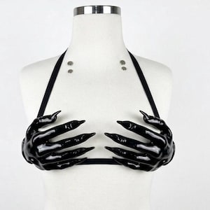 Könnte beinhalten: Ein schwarzes Neckholder-Top mit zwei glänzenden, geformten handförmigen Cups mit langen, spitzen Fingern. Das Kleidungsstück wird mit dünnen schwarzen Trägern befestigt und enthält zwei kleine silberfarbene Metallnieten im oberen Brustbereich.