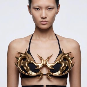 May include: A black bikini top featuring a metallic gold chest piece with an organic, spiked, and curved design. The gold ornament is structured with sharp points and smooth, rounded contours, layered over the black fabric of the bikini top.