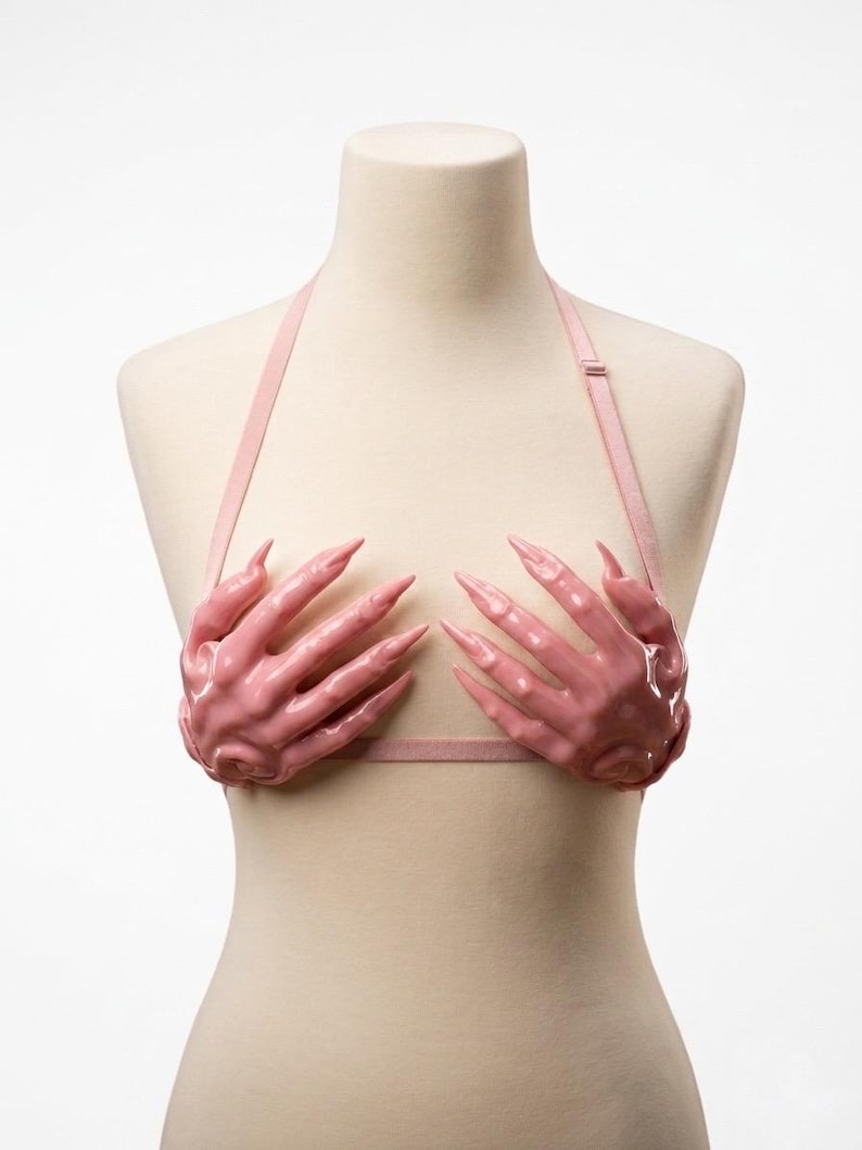 Peut inclure: Un soutien-gorge rose clair compos&eacute; de deux bonnets moul&eacute;s en forme de mains aux doigts longs et pointus. Le v&ecirc;tement comprend de fines bretelles r&eacute;glables assorties et une bande horizontale, le tout pr&eacute;sent&eacute; sur un buste de mannequin.