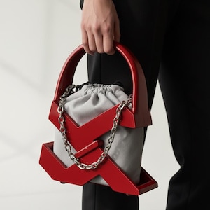 May include: A red geometric handbag with a silver chain strap and a gray drawstring pouch. The bag has a structured design with a curved handle. The bag is held by a person wearing black pants.