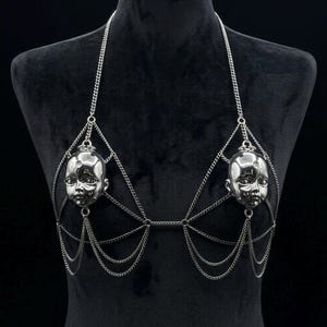 May include: A silver-toned body chain harness featuring two metallic doll head charms integrated into the design. The piece is constructed from delicate link chains that drape across the chest, connecting to the heads and forming a web-like structure.