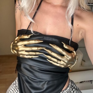 May include: A black faux leather halter top featuring two large, metallic gold-coloured sculpted hands with long, pointed fingers attached to the front. The top has thin black straps and a pointed hemline, paired with black and white chequered bottoms.