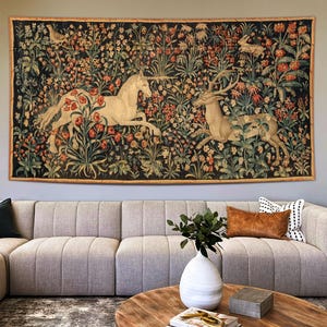 Puede incluir: Un gran tapiz que representa un unicornio y un ciervo en un entorno floral. La obra de arte presenta un fondo oscuro con flores y follaje coloridos. El tapiz está enmarcado y cuelga sobre un sofá seccional de color claro.