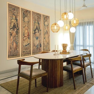 Op de afbeelding: Een eetkamer met een ovale tafel met een wit marmeren blad en een houten voet. Zes houten stoelen met grijze kussens omringen de tafel. Boven hangen hanglampen en aan de muur hangen vier ingelijste wandtapijten.