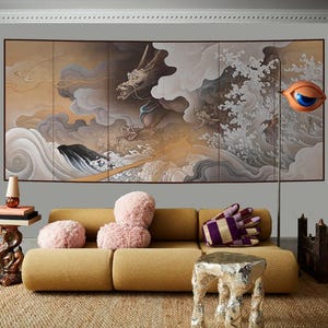Op de afbeelding: Een woonkamer met een mosterdkleurige bank, roze kussens en een handvormig kussen met een patroon. De muur toont een meerdelig kunstwerk van een draak in een stormachtige zee. Een oranje oogsculptuur en een zilveren bijzettafel zijn ook aanwezig.
