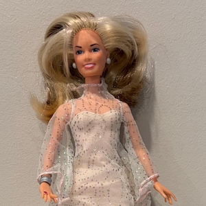 May include: A blonde doll wearing a light pink dress with silver glitter and a sheer overlay. Accessories include a black cat, a small orange book, and a rabbit on a black top hat. The doll has a smiling expression.