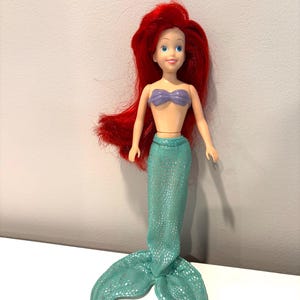 May include: A doll of a mermaid with bright red hair, blue eyes, and a purple seashell top. The doll has a turquoise tail with a shimmering pattern. The doll is standing upright.