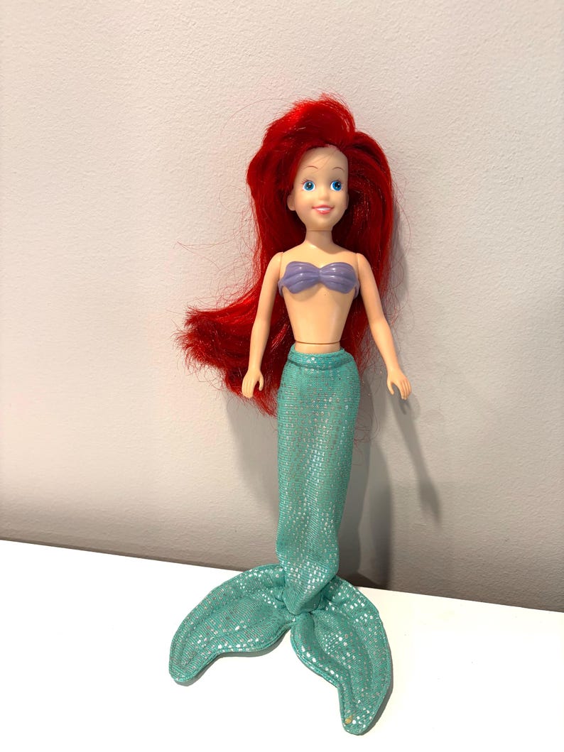 May include: A doll of a mermaid with bright red hair, blue eyes, and a purple seashell top. The doll has a turquoise tail with a shimmering pattern. The doll is standing upright.