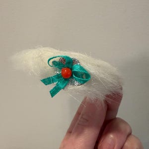 Puede incluir: Un accesorio para el cabello blanco y esponjoso con un lazo verde y una peque&ntilde;a cuenta roja. El lazo est&aacute; hecho de un material brillante y est&aacute; unido a una base plateada. El accesorio se sostiene en una mano.
