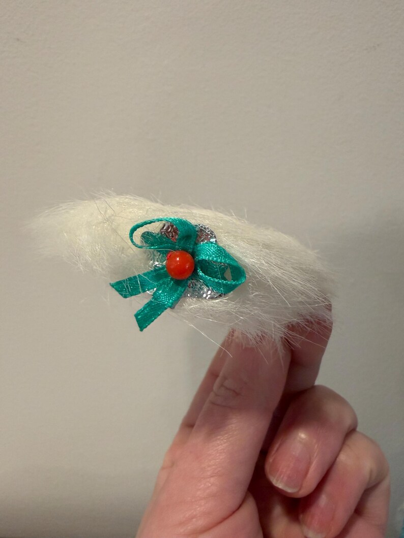 Puede incluir: Un accesorio para el cabello blanco y esponjoso con un lazo verde y una peque&ntilde;a cuenta roja. El lazo est&aacute; hecho de un material brillante y est&aacute; unido a una base plateada. El accesorio se sostiene en una mano.