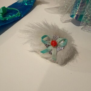 Puede incluir: Primer plano de un accesorio de mu&ntilde;eca, con un pomp&oacute;n blanco y esponjoso con una cinta turquesa y una peque&ntilde;a cuenta roja. Otros accesorios incluyen una diadema turquesa y una pieza azul con lentejuelas y ribete blanco.