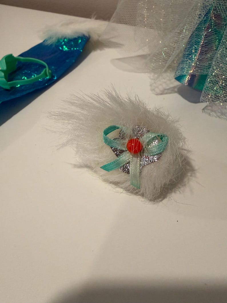 Puede incluir: Primer plano de un accesorio de mu&ntilde;eca, con un pomp&oacute;n blanco y esponjoso con una cinta turquesa y una peque&ntilde;a cuenta roja. Otros accesorios incluyen una diadema turquesa y una pieza azul con lentejuelas y ribete blanco.