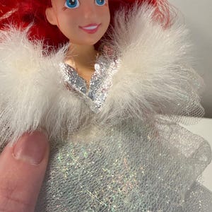 Puede incluir: Mu&ntilde;eca con cabello rojo y ojos azules, que lleva un chal blanco y esponjoso y un vestido plateado. La cara de la mu&ntilde;eca tiene una expresi&oacute;n sonriente. El vestido tiene una apariencia brillante e iridiscente.