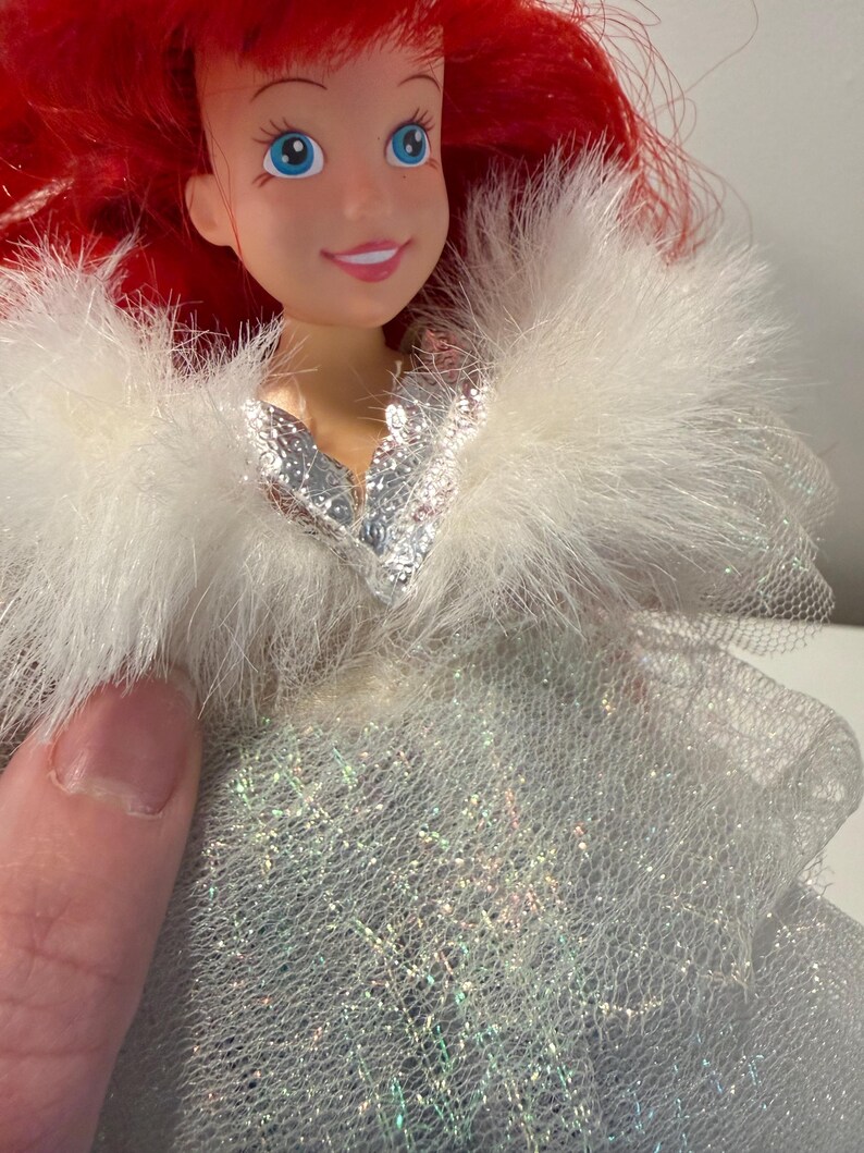 Puede incluir: Mu&ntilde;eca con cabello rojo y ojos azules, que lleva un chal blanco y esponjoso y un vestido plateado. La cara de la mu&ntilde;eca tiene una expresi&oacute;n sonriente. El vestido tiene una apariencia brillante e iridiscente.