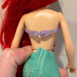 May include: A doll with red hair and a teal skirt. The doll's back is visible, showing a light purple top and the back of the doll's torso. The doll's hair is long and flowing.