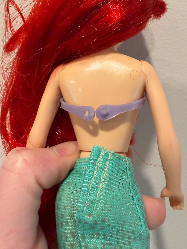 May include: A doll with red hair and a teal skirt. The doll's back is visible, showing a light purple top and the back of the doll's torso. The doll's hair is long and flowing.