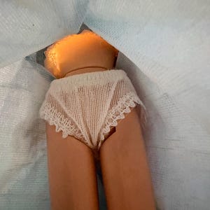 Puede incluir: Se ven el torso y las piernas de una mu&ntilde;eca, que lleva ropa interior color crema con ribete de encaje. La piel de la mu&ntilde;eca es de color bronceado claro. El pelo de la mu&ntilde;eca es de color naranja claro. La mu&ntilde;eca est&aacute; parcialmente cubierta por una tela azul claro.