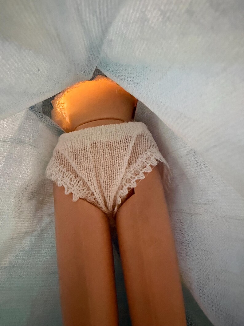 Puede incluir: Se ven el torso y las piernas de una mu&ntilde;eca, que lleva ropa interior color crema con ribete de encaje. La piel de la mu&ntilde;eca es de color bronceado claro. El pelo de la mu&ntilde;eca es de color naranja claro. La mu&ntilde;eca est&aacute; parcialmente cubierta por una tela azul claro.