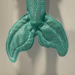 May include: A close-up of a teal mermaid tail made of fabric. The tail has a shimmering, silver dotted pattern. The tail is stuffed and has a stitched outline. The mermaid tail is a toy or decorative item.