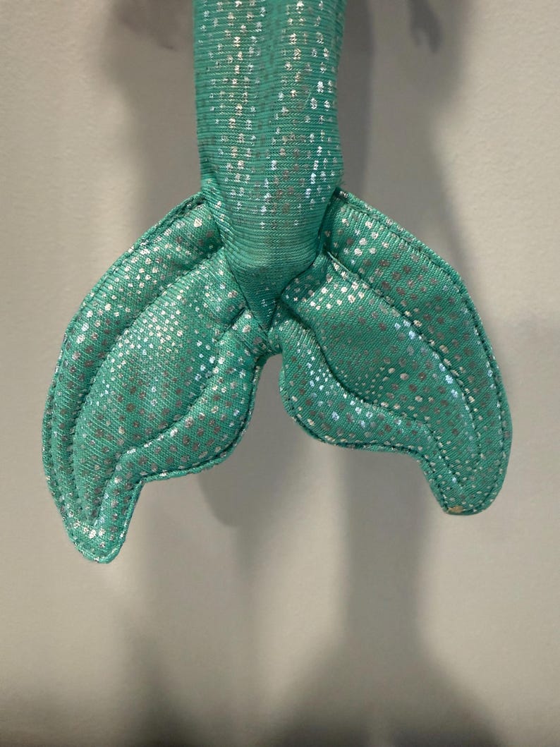 May include: A close-up of a teal mermaid tail made of fabric. The tail has a shimmering, silver dotted pattern. The tail is stuffed and has a stitched outline. The mermaid tail is a toy or decorative item.